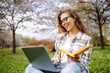 © maxbelchenko - Joyful woman sits with yellow notebook at table in blooming garden. Young woman works on laptop, holding notebook, on spring day. Concept of freelancing, relaxation, and weekends.