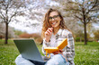 © maxbelchenko - Joyful woman sits with yellow notebook at table in blooming garden. Young woman works on laptop, holding notebook, on spring day. Concept of freelancing, relaxation, and weekends.