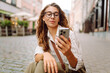 © maxbelchenko - Portrait of a young woman wearing glasses and holding a phone in a central square at sunset. A beautiful woman in stylish clothes texts on her phone on a sunny day. Concept of style and walking.