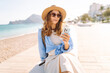 © maxbelchenko - A traveler wearing a hat and carrying backpack sits on bench near beach, holding phone. Young woman with a phone enjoys seascape at sunset. Conceptual ideas for tourism, recreation, and nature.