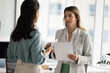 © fizkes - Two young adult businesswomen corporate employees colleagues team members meeting at modern office holding active business conversation discussing paperwork reviewing paper and electronic documents