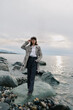 © SHOTPRIME STUDIO - Woman in coat and hat stands on rocky shore near water under cloudy sky. Casual fashion, autumn style, and outdoor mood with calm sea and natural landscape background.