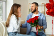 © Pixel-Shot - Young man giving bouquet of red rose flowers his surprised girlfriend and sitting on sofa in living room. Valentine's day celebration