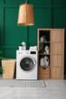 © Pixel-Shot - Washing machine, shelving unit, and laundry basket near green wall in room