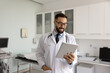 © fizkes - Virtual progress in medical care. Modern confident young doctor stand in contemporary examination room communicate with patient online review electronic records read digital chart on tablet with smile