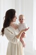 © New Africa - Smiling mother in dress holding her cute baby near white wall at home