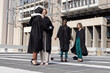 © wavebreak3 - Diverse female graduates hugging and walking on rooftop plaza in black gowns with mortarboards