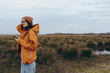 © SHOTPRIME STUDIO - Young woman lifestyle outdoors in autumn wearing orange jacket and knitted hat holding smartphone in natural landscape. Casual fashion and technology concept with moody sky and open field.