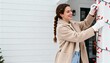 © Anna - Smiling woman hanging red Christmas lights on a white house pillar. Festive holiday home decoration concept. Female in beige coat and white gloves with copy space for text