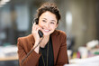 © Kowit - A smiling woman in a brown blazer talks on a corded telephone in a modern office setting.