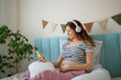 © wifesun - Pregnant woman lying on a bed, wearing wireless headphones and using a smartphone, enjoying music or meditation during pregnancy, supporting emotional and physical health