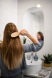© Zoran Jesic - Woman brushing long hair in bathroom mirror, practicing self-care