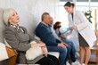 © JackF - Calm elderly woman sitting in clinic queue while nurse talks to other patients in background