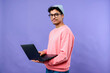 © pressmaster - Portrait of young adult Hispanic man wearing glasses looking at camera holding laptop standing against purple background, showing confident expression and casual posture