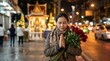 © jaturon - Hopeful Asian woman holding a bouquet of red roses and praying with closed eyes at a city shrine on a street at night.