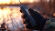 © Jdgkxbnc - gloved hand in camouflage holding a handheld radio with antenna by a marshy shoreline at sunset, warm golden light and a calm focused mood