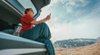 © olezzo - Woman in red t-shirt using smartphone while sitting in an open car trunk during a mountain road trip. Solo traveler checking map or social media with scenic mountain view.