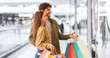 © Prostock-studio - A couple stands inside a mall holding colorful shopping bags. They are looking at store displays and smiling, enjoying their time shopping together.