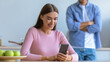 © Prostock-studio - A woman is sitting at a kitchen table, looking at her smartphone and smiling. A man stands behind her with crossed arms, observing her. The setting has green apples on the table.