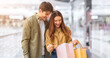 © Prostock-studio - A man and a woman stand close in a shopping mall. They look happy as they open shopping bags together. The man smiles while the woman shows excitement about the items inside the bags.