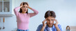 © Prostock-studio - A woman stands with her hands on her head, showing signs of frustration. A man sits nearby, holding his temples, indicating stress. The room appears to be a kitchen.