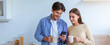 © Prostock-studio - A man and a woman stand together in a kitchen. They hold cups and smile while looking at a smartphone. It is morning, and the kitchen has a light atmosphere with various items visible.