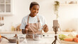 © Prostock-studio - A woman is cooking in a modern kitchen. She smiles while adding ingredients to a pot and holds a spoon in her hand. A camera is set up to record her cooking process.
