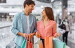 © Prostock-studio - Two people are smiling at each other while holding shopping bags. They are in a mall with many shoppers in the background. The scene shows a lively atmosphere and happy moments.