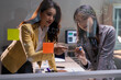 © Tj - Two young asian businesswomen discussing and writing ideas on a glass board with sticky notes in a modern office setup, collaborating on a challenging business project