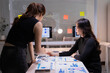© Tj - Two businesswomen collaborating on a strategy, reviewing market data sheets and financial charts while brainstorming ideas on a whiteboard in a modern office environment