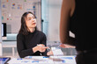 © Tj - Young businesswoman pointing at a calculator and engaging in a discussion with a colleague, analyzing financial data and charts spread across the desk during a corporate meeting in an office setting