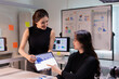 © Tj - Two businesswomen are discussing analytical data on a digital tablet, pointing at graphs during a collaborative meeting in a modern office with charts on a whiteboard