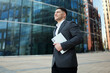 © muse studio - Young man walks confidently outside a modern building holding a laptop during daytime