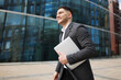 © muse studio - Businessman walks outside a modern office building while carrying a laptop during the day