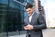 © muse studio - Man in a suit smiles while using a smartphone outside a modern building in the city