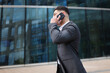 © muse studio - Young man talks on phone while walking outside a modern building in a city setting
