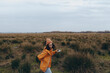 © SHOTPRIME STUDIO - Woman lifestyle concept with a young woman in an orange jacket and beanie walking outdoors in a field with tall grass under a cloudy sky. Casual autumn fashion and nature exploration.