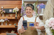 © Ekkasit A Siam - Middle-aged Asian female florist in apron holding credit card and card reader in flower shop, smiling for contactless payment service concept with fresh flowers and professional retail sales.