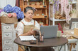 © Ekkasit A Siam - Senior asian woman florist reading notes while sitting beside laptop showing confused or tired face surrounded by flowers and books reflecting difficulties running small floral business