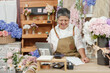 © Ekkasit A Siam - Senior asian florist smiling and looking at mobile phone while sitting at flower shop counter showing joyful emotion after selling flower products online colorful arrangements visible behind