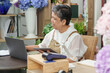 © Ekkasit A Siam - asian senior florist talking and pointing at laptop screen while holding tablet seated at flower shop counter with stacked tools bouquet and plants nearby during online business management