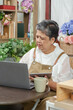 © Ekkasit A Siam - Asian senior florist sitting in cozy flower shop working with laptop and tablet smiling confidently expressing happiness from successful online business and growing digital sales
