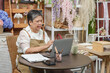 © Ekkasit A Siam - asian senior female florist wearing apron sitting at table checking inventory using laptop in flower shop with clipboard smartphone and pencil holder surrounded by colorful floral arrangements
