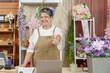 © Ekkasit A Siam - Smiling senior Asian florist wearing apron pointing forward in floral studio standing behind desk with laptop clipboard and flower arrangements showing confidence and warmth