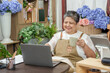 © Ekkasit A Siam - asian senior florist laughing holding cup while sitting at flower shop desk with open notebook and laptop feeling accomplished with business sales results and joyful digital workspace