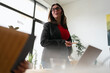 © Studio Marmellata - A woman in glasses and a blazer holds a pencil while speaking to colleagues in a modern office setting with laptops and plants.