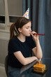 © Volha - Sad looking teenage girl wearing party hat blowing party horn and celebrating birthday online using laptop, with slice of birthday cake and lit candle in front of her. Vertical photo