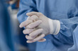 © wedmoments.stock - Medical professional in blue surgical attire with gloved hands clasped together, preparing for a procedure in a clinical environment with blurred background elements