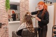 © Renata Hamuda - Smiling redhead Hairdresser curling hair of female client using iron in salon