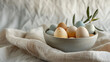 © Anom S. - Naturally colored pastel farm eggs displayed in a rustic bowl on soft linen with an olive branch, evoking spring's fresh, organic charm